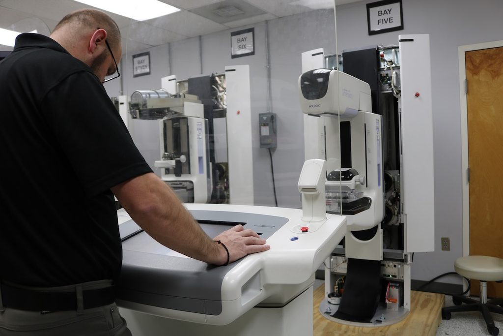 Technician working at a Hologic acquisition workstation in front of Selenia Dimensions mammography systems with their covers opened for service in numbered repair bays.